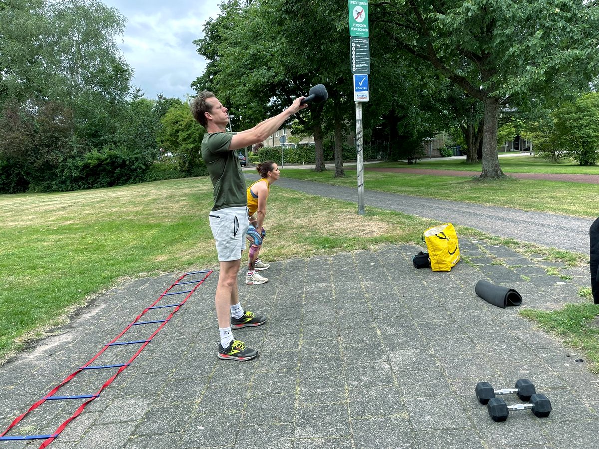 Twee mensen trainen met kettlebells op een verhard pleintje met een ladder op de grond