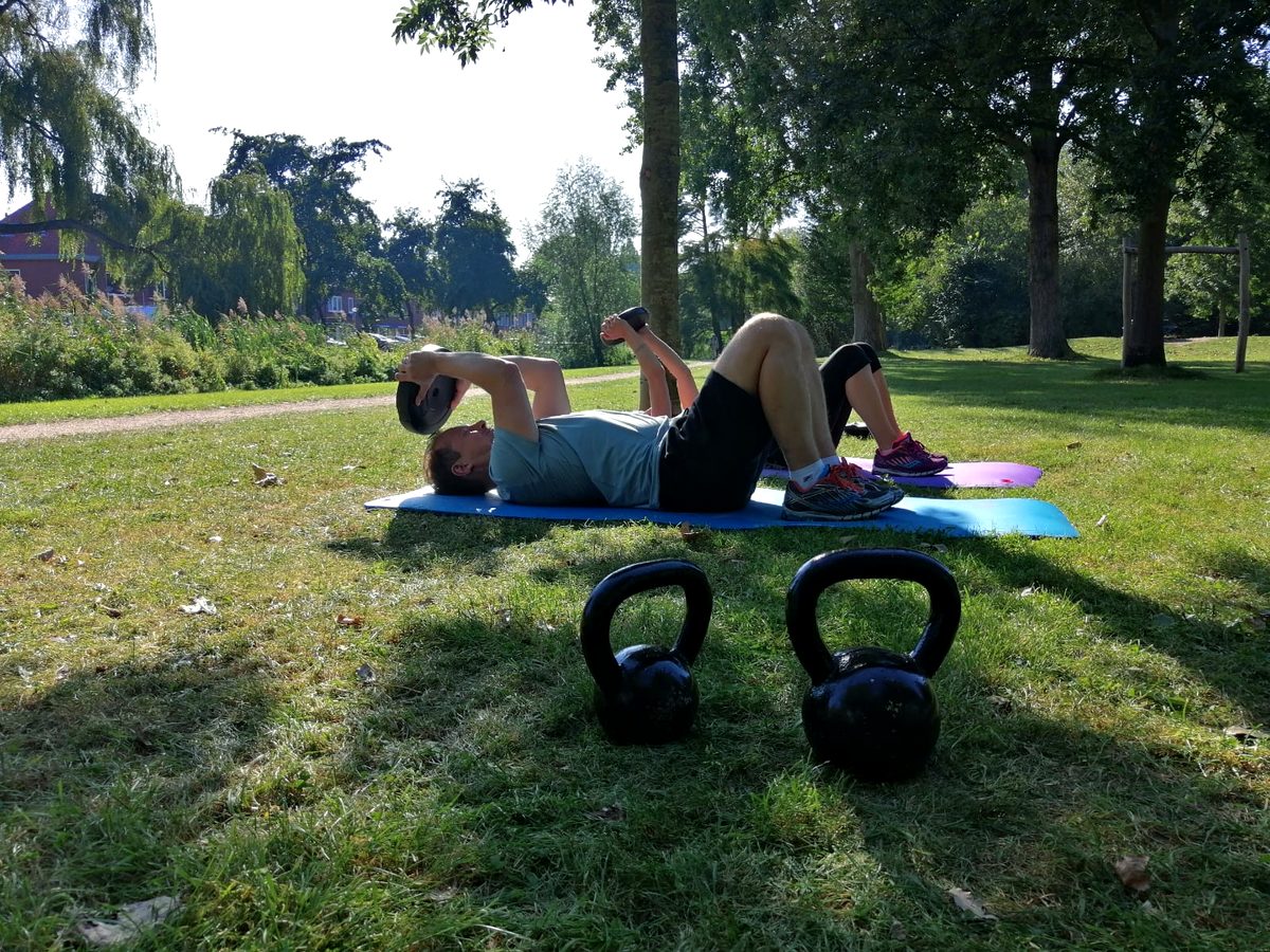 Twee mensen doen buikspieroefeningen naast elkaar op matten in het gras, met kettlebells ernaast